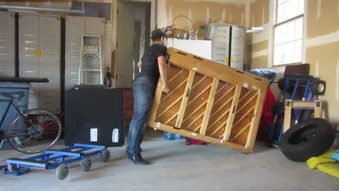 a man tilts a crated upright piano onto a wheeled dolly in a garage