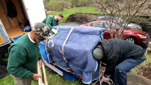 three movers loading a wrapped upright piano onto a board by the truck, face visible