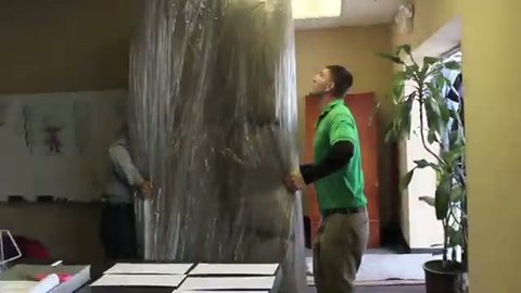 two movers carrying a shrink-wrapped sofa indoors, face visible